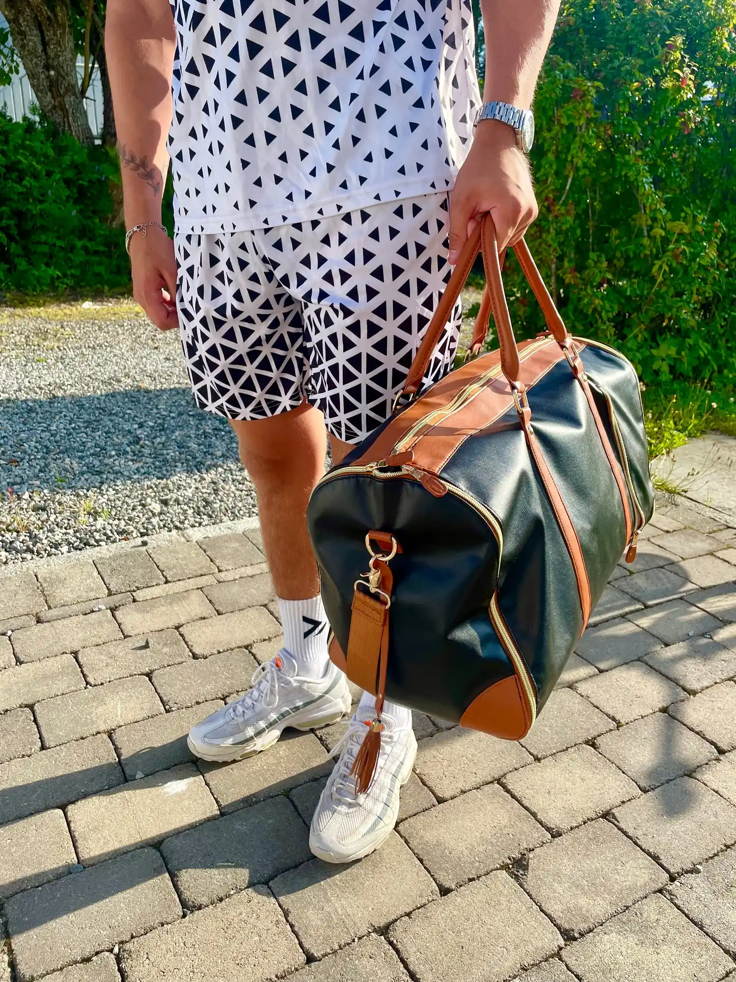 Svart reisebag med brune detaljer og en dusk bæres av en person i mønstrede klær utendørs, på brostein med busker i bakgrunnen.