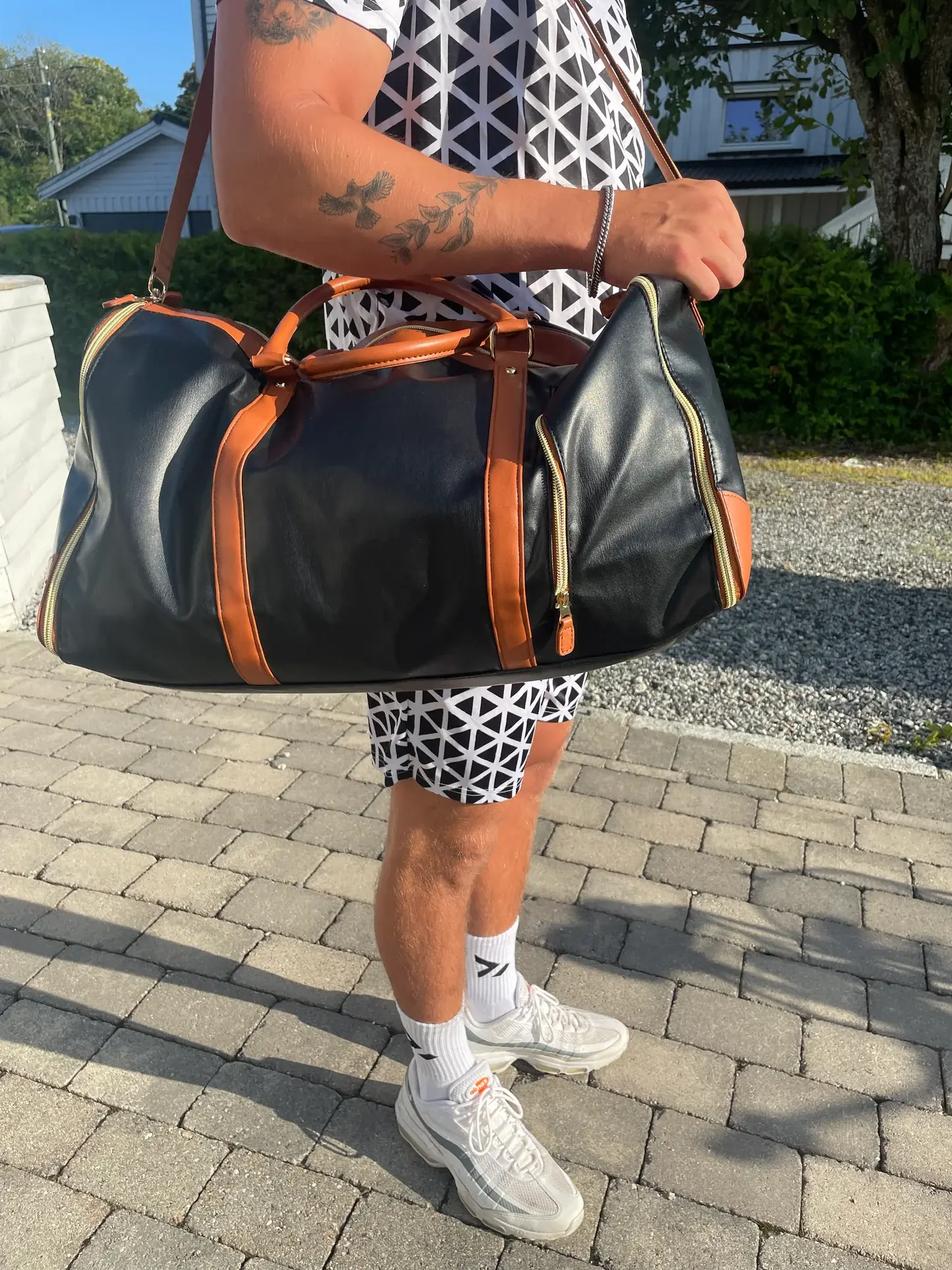 Svart reisebag med brune detaljer bæres over skulderen av en person i mønstrede klær utendørs, på brostein med busker og en bolig i bakgrunnen.
