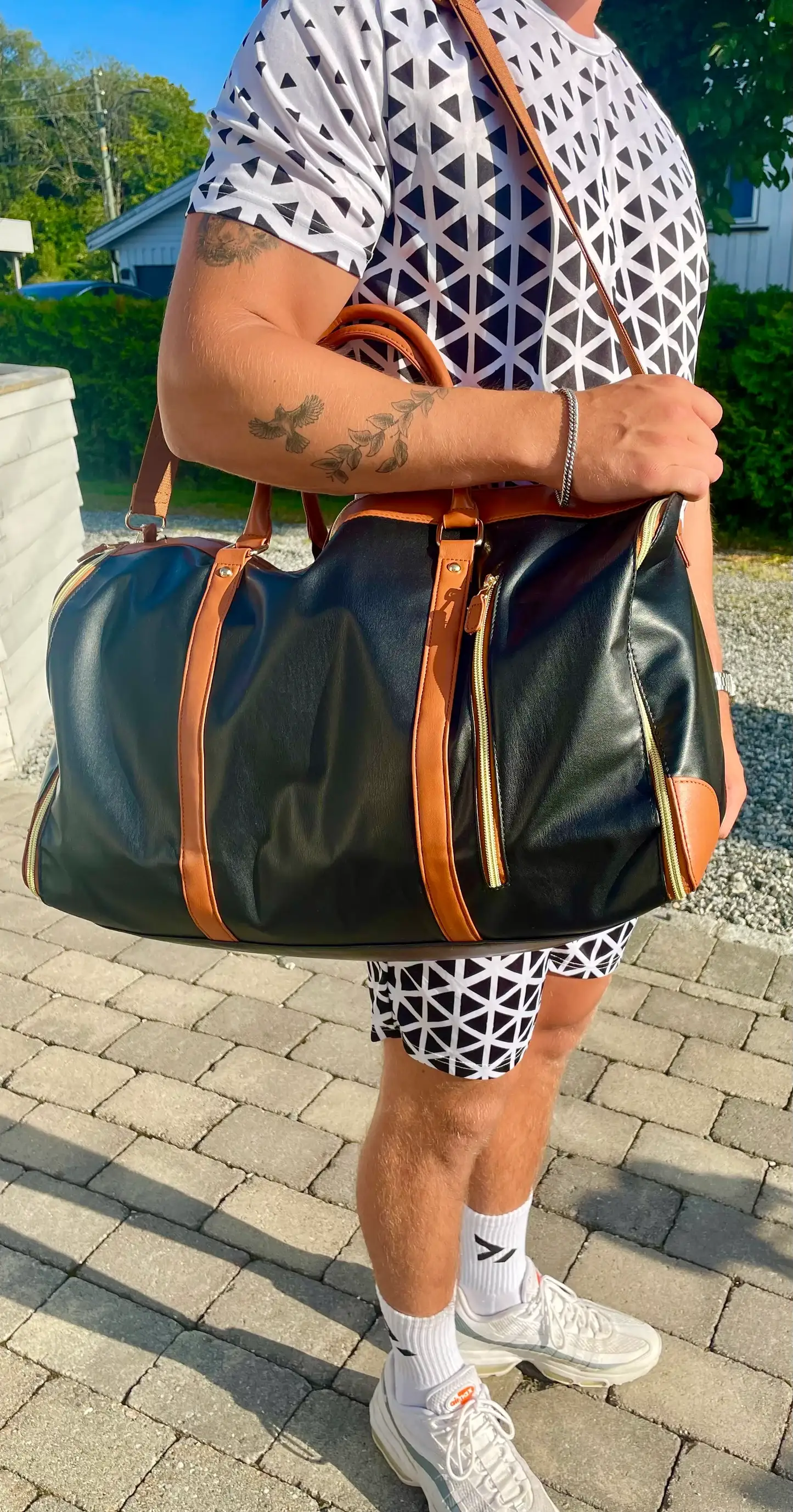 Svart reisebag med brune detaljer bæres av en person i mønstrede klær utendørs på brostein, med en lys bolig og grønne busker i bakgrunnen.