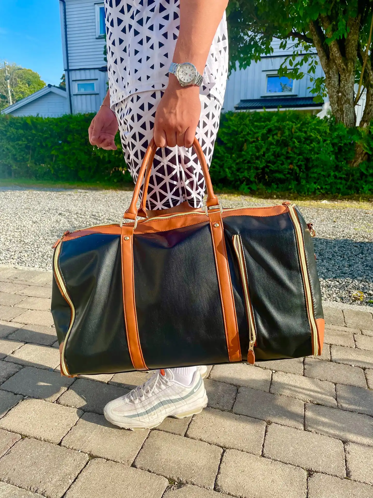 Svart reisebag med brune detaljer holdes i hånd av en person i mønstrede klær, utendørs på brostein med en grønn hekk og lys bolig i bakgrunnen.