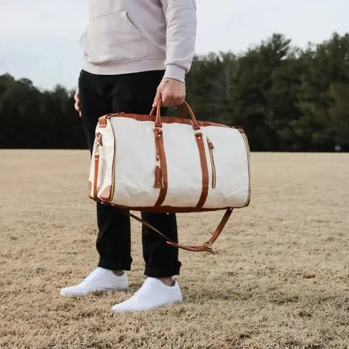 Hvit reisebag med brune detaljer bæres av en person i svart bukse og hettegenser, stående på en gressplen med trær i bakgrunnen.