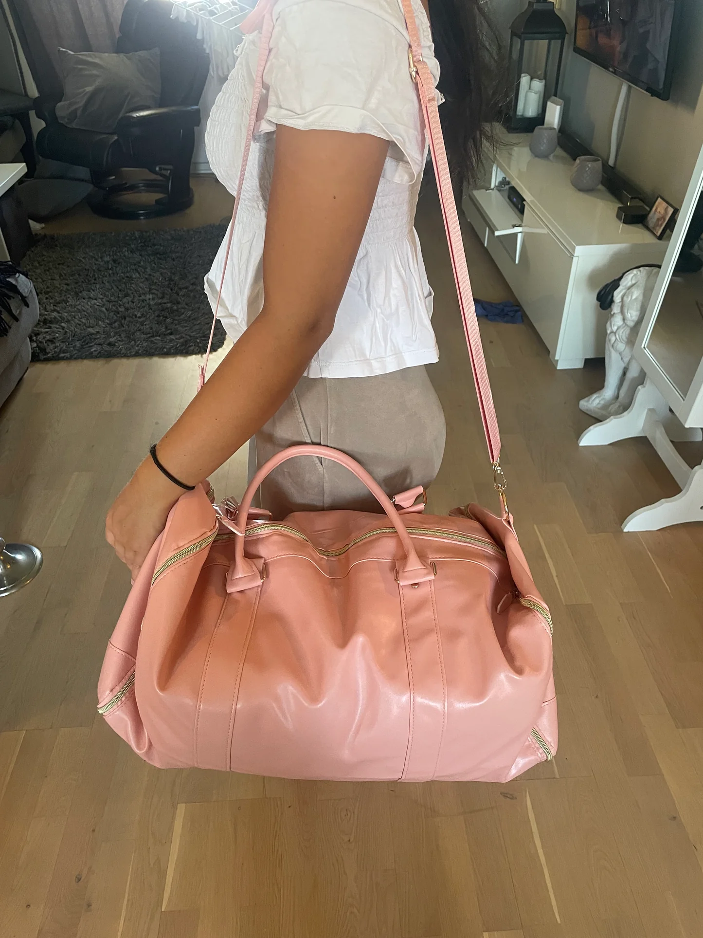 Rosa reisebag med gyldne detaljer bæres av en person i et stueinteriør med lyse tregulv og moderne møbler.